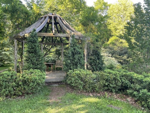 A rustic gazebo surrounded by lush greenery and shrubs in a serene garden setting.