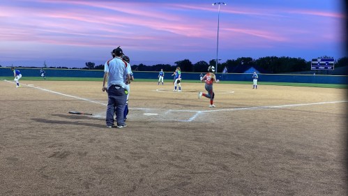 A softball player runs toward home plate during a game at sunset, with teammates and coaches in the background.
