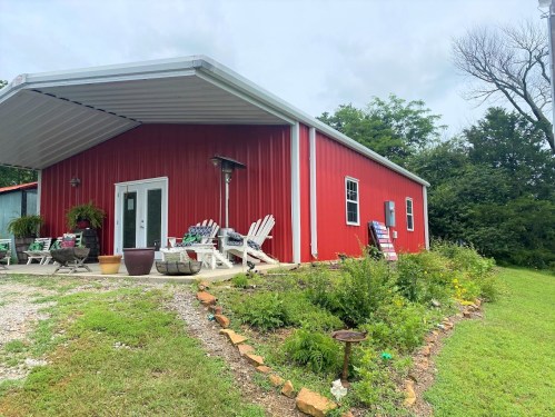 A red metal building with a covered porch, surrounded by greenery and a flower garden, under a cloudy sky.
