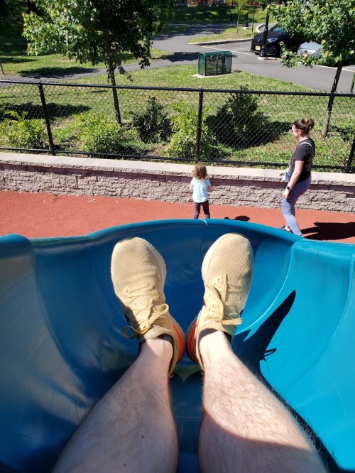 View from a slide, showing feet in sneakers, with a child and adult in the background at a playground.