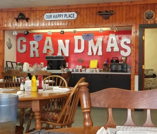 Interior of a cozy diner with a red wall displaying "GRANDMAS" and a sign that says "OUR HAPPY PLACE." Tables set for dining.