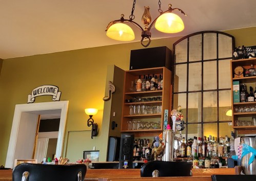 A cozy bar interior featuring a wooden counter, shelves of drinks, and warm lighting with a "Welcome" sign.