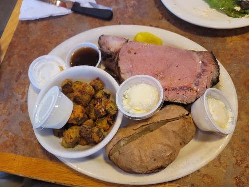 A plate with sliced roast beef, baked potato, fried okra, and dipping sauces, garnished with a pickle.