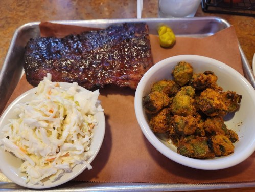 A tray with a piece of glazed rib, coleslaw, and fried okra, accompanied by a small dollop of mustard.