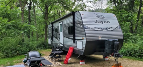 A Jayco travel trailer parked in a wooded area, with camping chairs and a grill set up nearby.