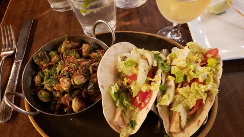 Two tacos filled with lettuce, tomatoes, and fish, served with a side of Brussels sprouts and a drink.