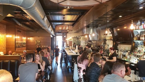 A bustling restaurant interior with patrons at tables and a bar, featuring warm lighting and a stylish design.