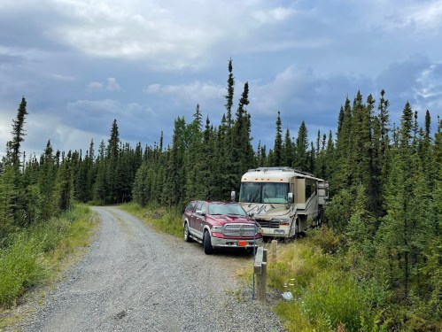 An RV and a red truck parked on a gravel road surrounded by tall green trees under a cloudy sky.