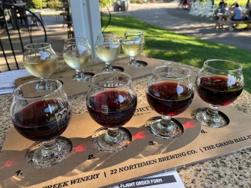 A tasting flight of wines and beers in various glasses on a table, with a sunny outdoor setting in the background.