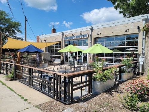 Outdoor seating area of a brewery with colorful umbrellas, surrounded by plants and flowers under a clear blue sky.
