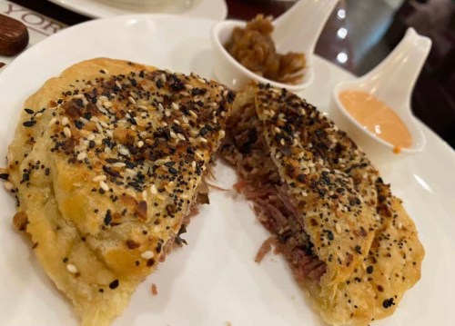 A plate featuring a split, flaky pastry filled with meat, topped with sesame seeds, and served with two sauces.