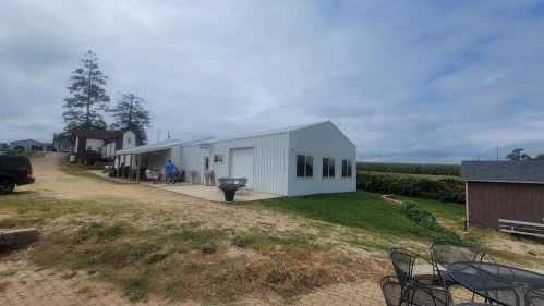 A white metal building on a farm, surrounded by greenery and a gravel path, with outdoor seating nearby.