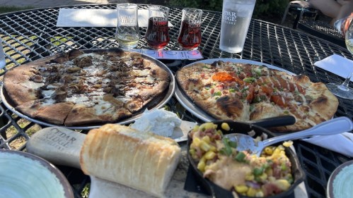 A table with two pizzas, a bread roll, and a skillet of corn salsa, alongside drinks at a winery.