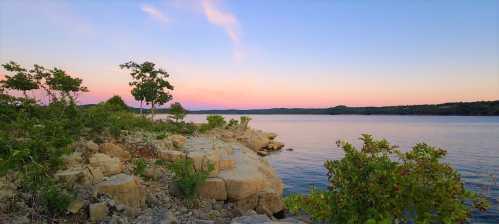 A serene lakeside view at sunset, featuring rocky shores and lush greenery under a colorful sky.