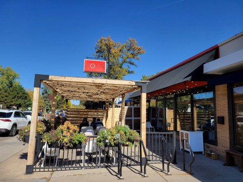 Outdoor dining area of a restaurant with a wooden pergola, surrounded by flowers, under a clear blue sky.