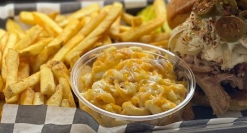 A plate featuring golden fries, a cup of cheesy macaroni, and a sandwich topped with jalapeños and coleslaw.