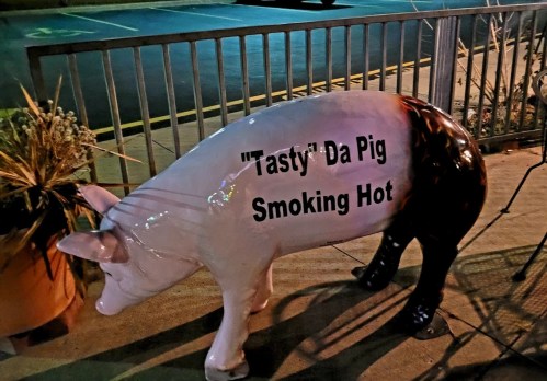 A painted pig statue with the text "Tasty! Da Pig Smoking Hot" displayed on its side, positioned outdoors.
