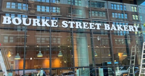 Signage for Bourke Street Bakery on a glass storefront, with a ladder nearby and reflections of buildings.