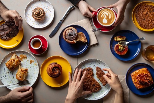 A top-down view of a table with various pastries, coffee, and hands reaching for food on colorful plates.