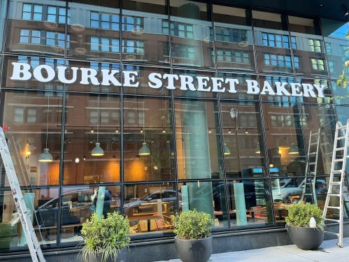Exterior of Bourke Street Bakery with large signage and potted plants, reflecting a city street in the background.