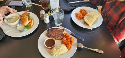 Three plates of food on a table: a burger, a taco, and a meat dish with mashed potatoes and carrots, with drinks nearby.