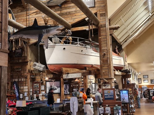 A large boat hangs from the ceiling in a rustic store, with a shark model and shoppers browsing below.