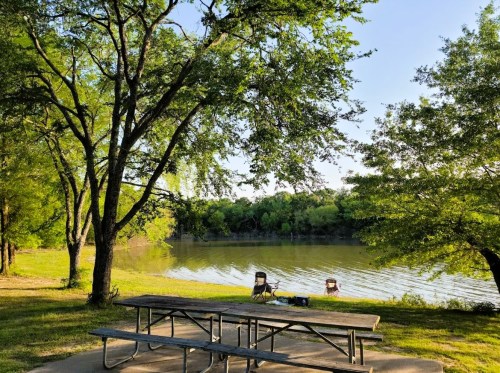 A serene lakeside scene with a picnic table and two chairs under trees, reflecting calm water and greenery.