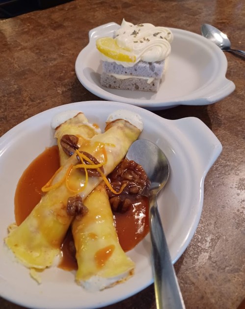A plate with three rolled crepes topped with sauce and nuts, alongside a slice of dessert with cream and lemon garnish.
