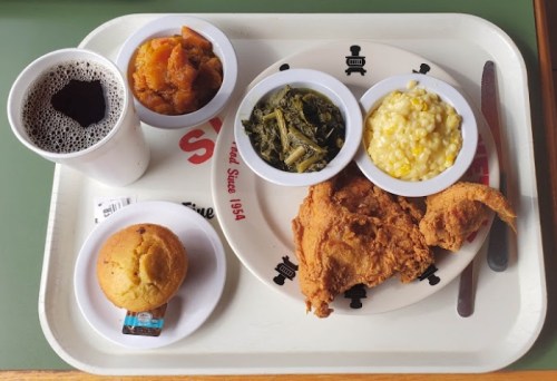 A tray with fried chicken, collard greens, corn, sweet potatoes, a muffin, and a cup of soda.