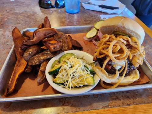 A tray with a burger topped with onion rings, sweet potato fries, and a side of zucchini with cheese.