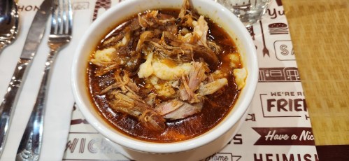 A bowl of savory soup with shredded chicken, spices, and chunks of cauliflower, served on a patterned table.