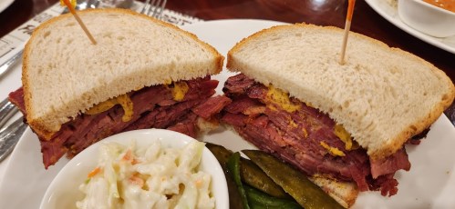 A close-up of a sandwich with layers of meat and mustard, served with coleslaw and pickles on the side.