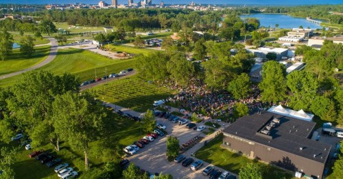 Aerial view of a large crowd gathered in a park near a river, with cars parked and city skyline in the background.