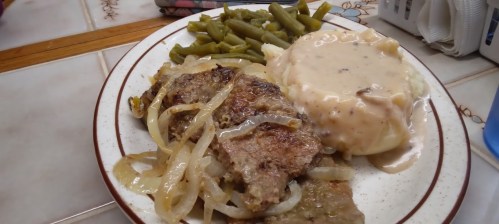 A plate of meatloaf with onions, green beans, and mashed potatoes topped with gravy.