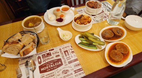 A table spread with various dishes, including bread, pickles, and bowls of soup and desserts, set on a patterned tablecloth.