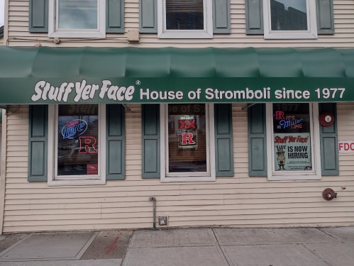 Sign for "Stuff Yer Face," a Stromboli restaurant since 1977, with neon lights and a hiring notice in the windows.