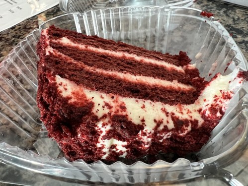 A slice of red velvet cake with cream cheese frosting in a clear plastic container.