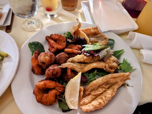A plate of fried seafood including shrimp, fish, and hushpuppies, garnished with lemon and served on a bed of greens.