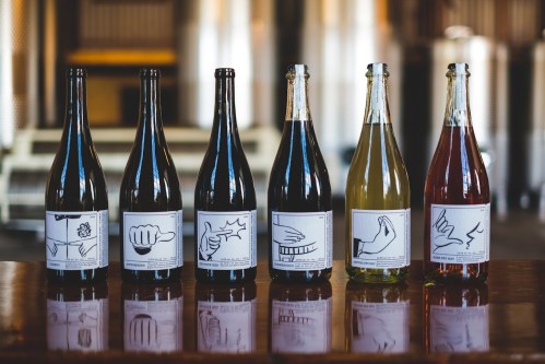 Six bottles of wine with unique illustrated labels, displayed on a wooden table in a bright, industrial setting.