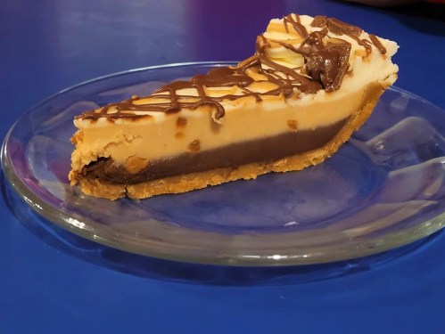 A slice of layered dessert on a glass plate, featuring chocolate and caramel layers with a drizzle of chocolate on top.