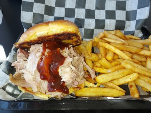 A pulled pork sandwich with barbecue sauce on a bun, served with a side of golden French fries in a checkered tray.