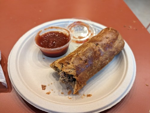 A partially eaten pastry on a paper plate, accompanied by two small cups of salsa.