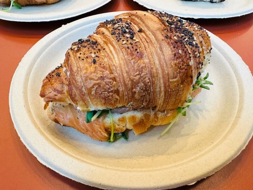 A croissant sandwich filled with salmon and greens, served on a biodegradable plate.