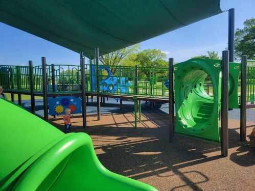 A colorful playground with green slides and climbing structures under a shaded area, surrounded by trees.