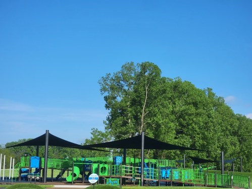 A colorful playground with green equipment and shade sails under a clear blue sky, surrounded by trees.