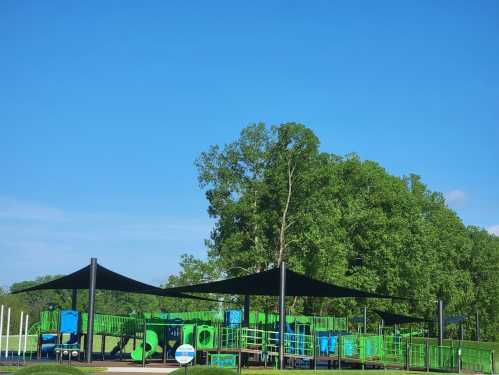 A colorful playground with green equipment and shade sails under a clear blue sky, surrounded by trees.