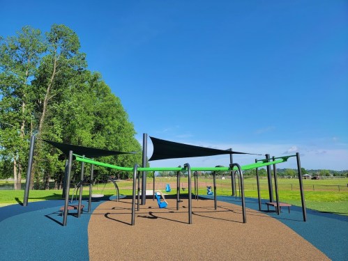 A colorful playground with green equipment and shade sails, set in a grassy area under a clear blue sky.