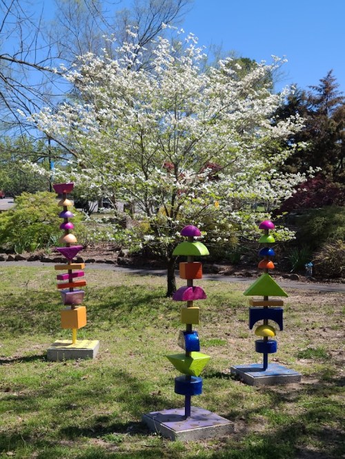 Colorful abstract sculptures stand in a garden with a flowering tree and greenery in the background under a clear blue sky.