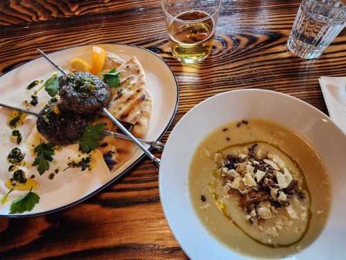 A plate with skewered meat, flatbread, and herbs, alongside a bowl of soup drizzled with olive oil and topped with cheese.