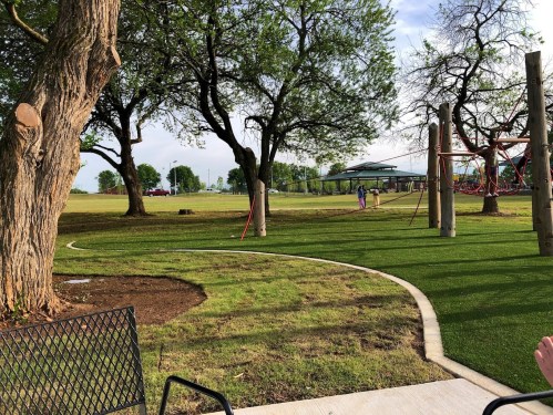A park scene with green grass, trees, and a playground in the background, viewed from a patio area.